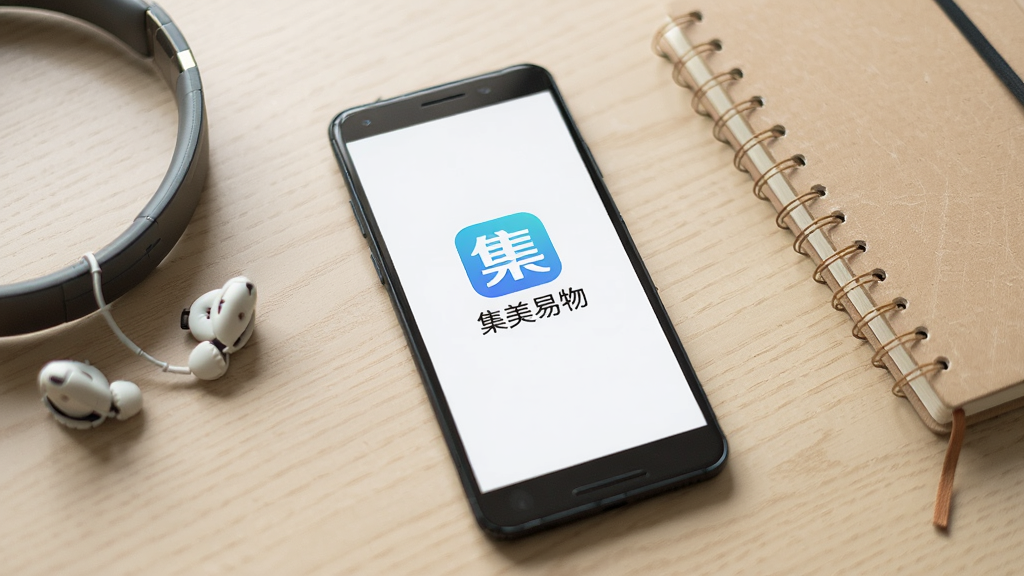 一部安卓手机放置在桌面上,屏幕显示集美易物的启动页面,页面中央为应用图标和名称“集美易物”;手机旁边放着一副耳机和一本笔记本,背景为浅木色桌面;整体光线柔和,构图简洁,画面居中,细节清晰,画质干净,无多余元素。