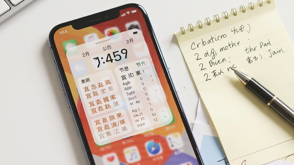 手机屏幕显示中脉黄历万年历桌面小组件效果,小组件展示当日公历农历日期、宜忌摘要与节气信息,背景为手机主屏壁纸。手机旁放置一张手写便签与一支笔。风格为生活化场景展示,光线明亮,构图自然,画质干净,无多余元素。