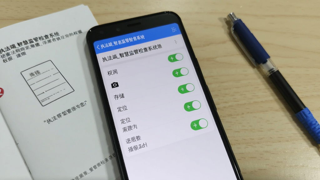 一部安卓手机显示“执法端_智慧监管检查系统”的权限设置页面,页面上相机、存储、定位等权限开关均处于开启状态,手机旁边放着说明书和笔,整体风格简洁实用,色调清爽,画质清晰,无多余元素