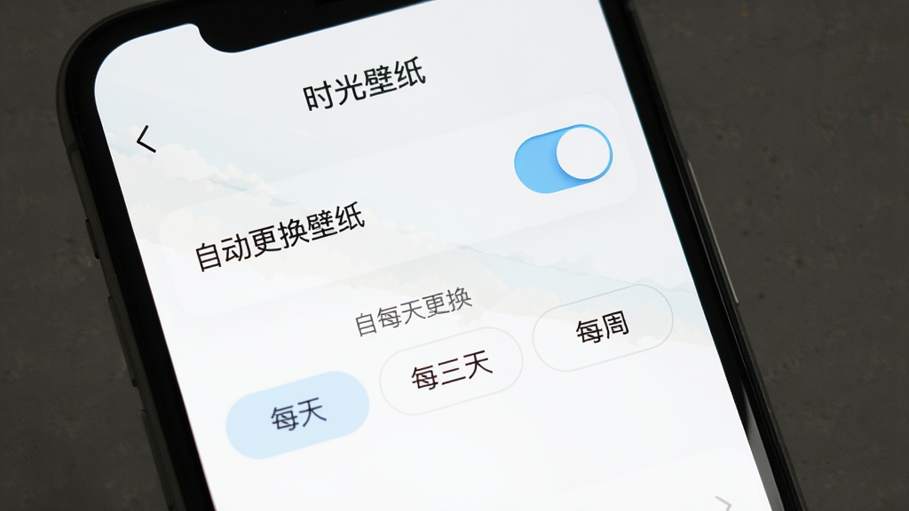 手机屏幕显示“时光壁纸”设置页面,页面上方有“自动更换壁纸”开关按钮,下方有更换频率选项如“每天”“每三天”“每周”,界面风格极简,白色背景配淡蓝色开关按钮,排版清晰,无多余图标,高清画质,文字“自动更换”清晰可见。