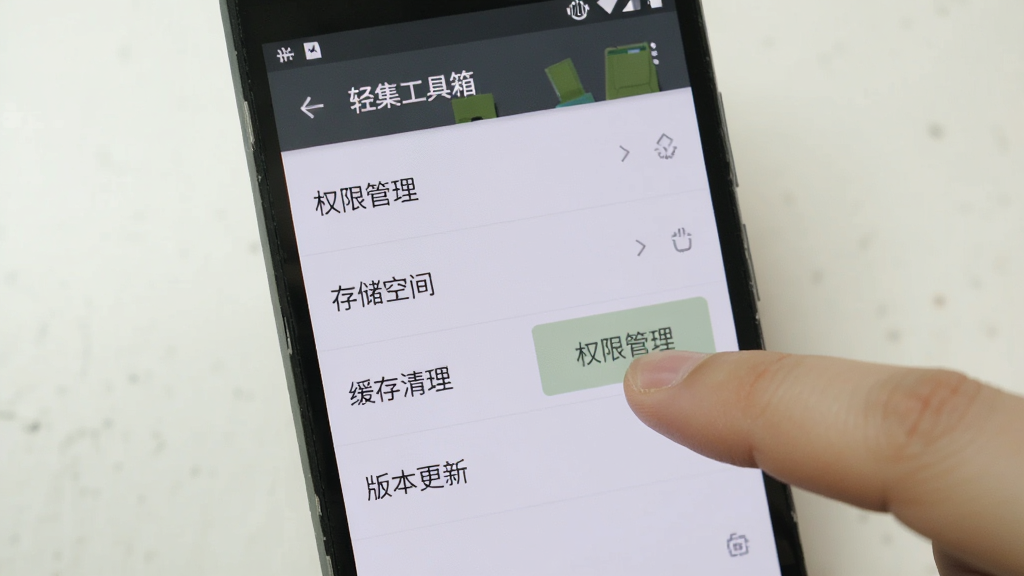 一部安卓手机屏幕显示“轻集工具箱”的设置页面,页面中有“权限管理”“存储空间”“缓存清理”“版本更新”等选项,画面右侧有一只手指正在点击“权限管理”按钮,背景为浅色,风格写实,画质清晰,干净无多余元素