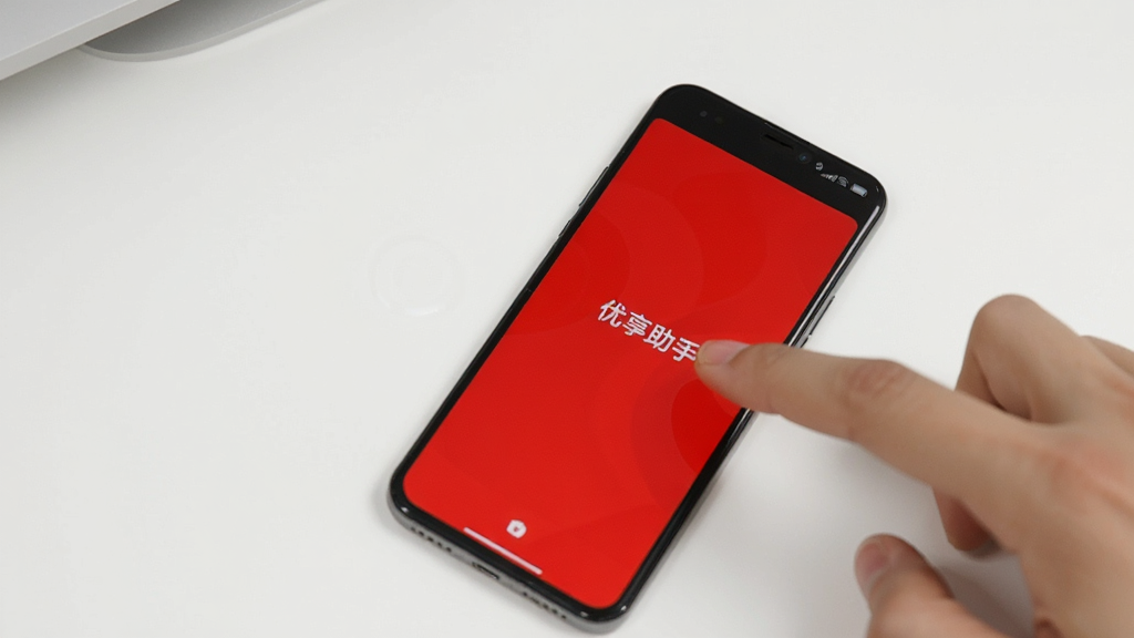 一部手机显示“优享助手”屏幕测试界面,屏幕呈现纯红色,手机旁边有一只手正在触摸屏幕,背景为白色桌面,画面简洁明亮,画质清晰