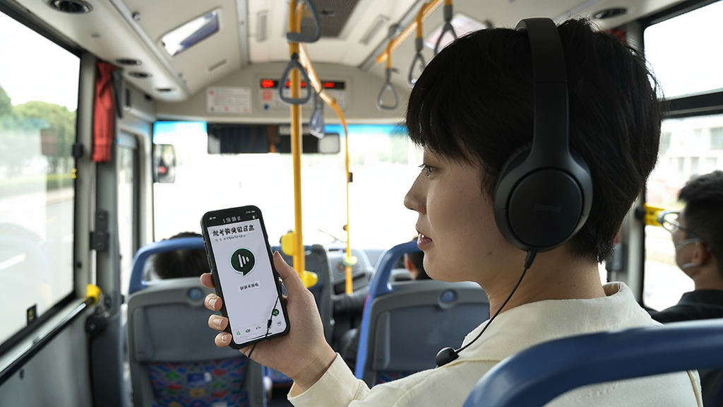 一位年轻学员戴着耳机坐在公交车上,手持手机使用“驾考宝典语音盒”听题,手机屏幕显示语音播报界面,周围环境为明亮的车厢内部,风格写实温馨,细节丰富,画质清晰,干净无多余元素