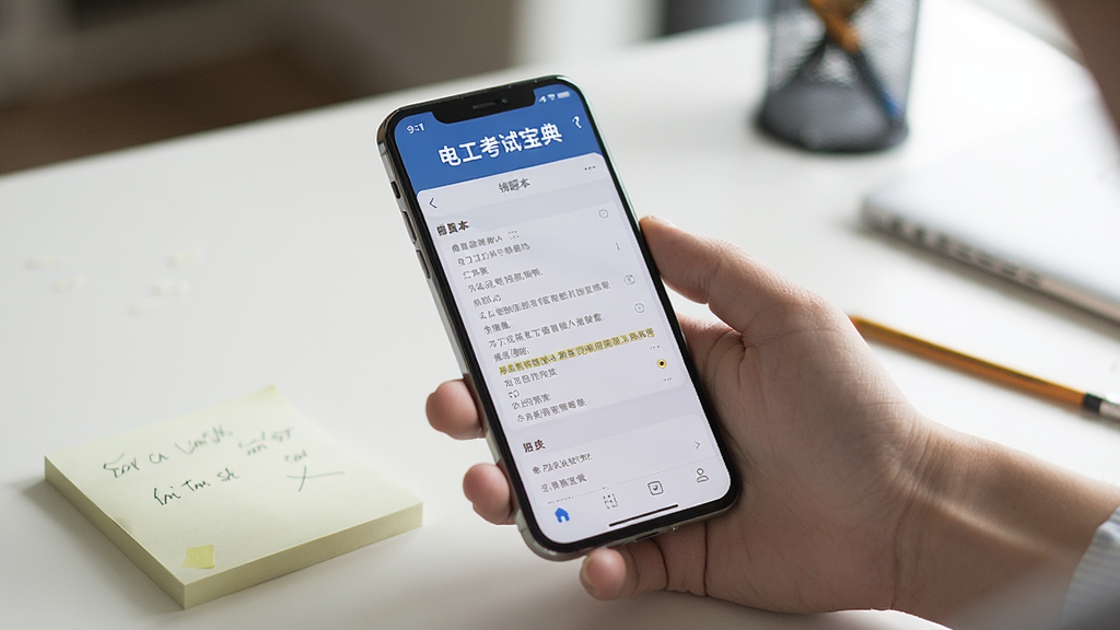 一张手机使用场景图,画面中一只手握着手机,屏幕显示“电工考试宝典”的错题本页面,旁边有便签纸和铅笔,桌面整洁,背景虚化,风格生活化,画质清晰,突出手机屏幕内容