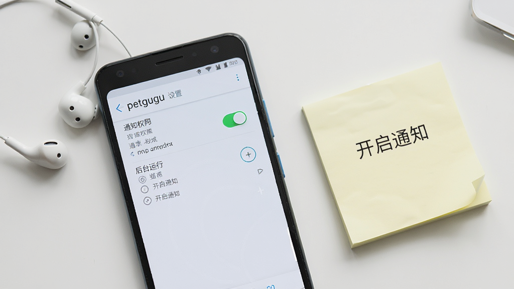 一部安卓手机屏幕显示petgugu设置页面,页面中有通知权限开关和后台运行选项,手机旁边放着耳机和便签纸,便签上写着“开启通知”,桌面整洁,正面视角构图,风格简洁实用,细节清晰,画质干净明亮,无多余元素