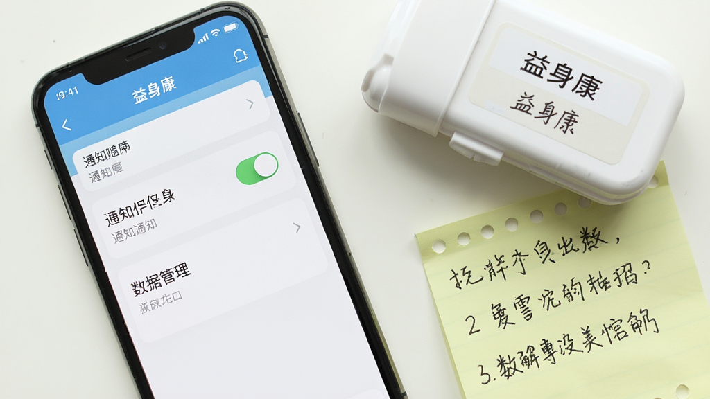 画面主体是一部手机屏幕显示“益身康”应用的设置页面,页面上有通知权限开关和数据管理选项;手机旁边放着一个小药盒和一张便签,便签上写着提醒事项;布局为桌面平铺俯拍,手机位于画面左侧,物品错落摆放;排版自然随意;风格为生活记录场景,色调明亮;细节包括屏幕设置项清晰、药盒标签可见、便签字迹自然;画质清晰,干净,无多余元素。