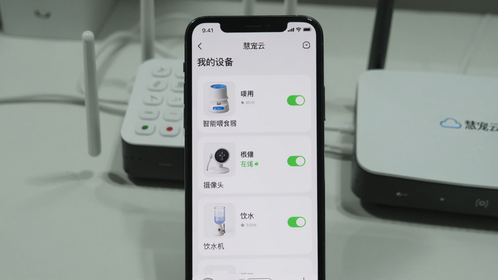画面主体为一部手机竖屏显示“慧宠云”APP的“我的设备”页面,列表中有智能喂食器、摄像头、饮水机三个设备卡片,每个卡片右侧有开关按钮,其中一个显示在线状态。布局为手机居中,背景为浅灰色桌面,旁边放着一个WiFi路由器。排版整洁,设备图标清晰。风格为写实摄影风格,光线明亮,细节体现屏幕内的绿色状态圆点。画质干净无多余元素。