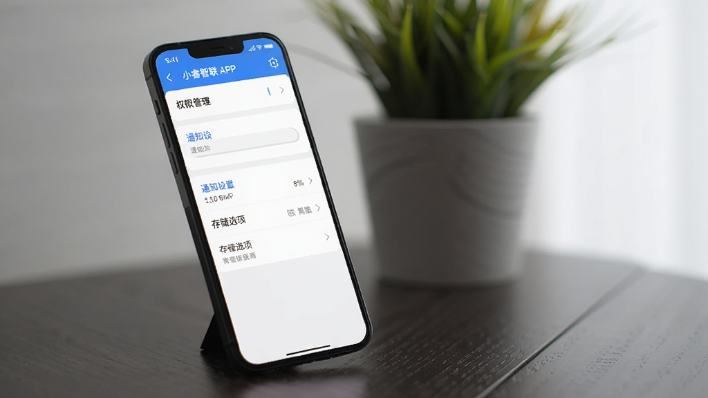 画面主体为一部智能手机屏幕展示小睿智联APP的设置页面,页面中显示权限管理、通知设置、存储选项等菜单列表。手机倾斜放置在深色木纹桌面上,屏幕亮起,界面清晰可见,背景有一盆绿植虚化,整体风格简洁自然,光线柔和,细节丰富,画质干净。