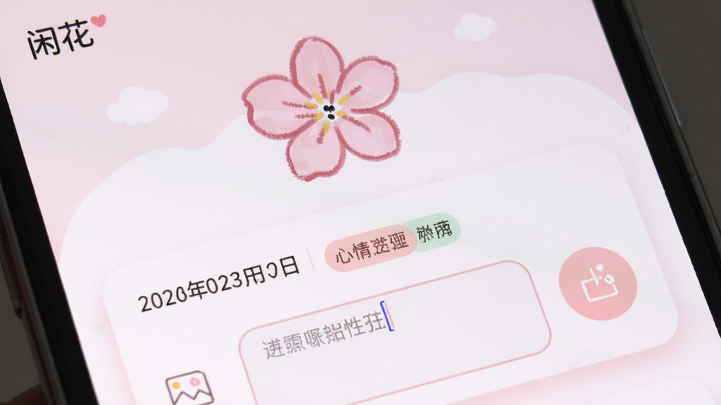 手机屏幕特写,显示“闲花”App的心情日记界面,界面以淡粉色和白色为主,上方有一朵手绘风格的粉色花朵,下方是日期和心情标签,旁边有文字输入框和照片添加按钮,整体排版简洁,风格清新可爱,细节包括柔和阴影和圆角边框,画质细腻干净,无多余元素