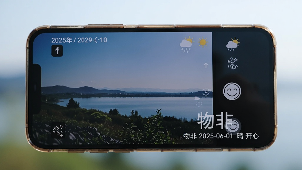 主体:一部手机横屏拍摄一张带有“物非”水印的照片,照片角落显示日期、天气和心情图标。布局:手机居中,背景为模糊的户外风景。排版:水印文字清晰,位于照片右下角。风格:纪实风格,色彩自然。细节:水印内容为“物非 2025-06-01 晴 开心”。画质:高清,干净,无多余元素。