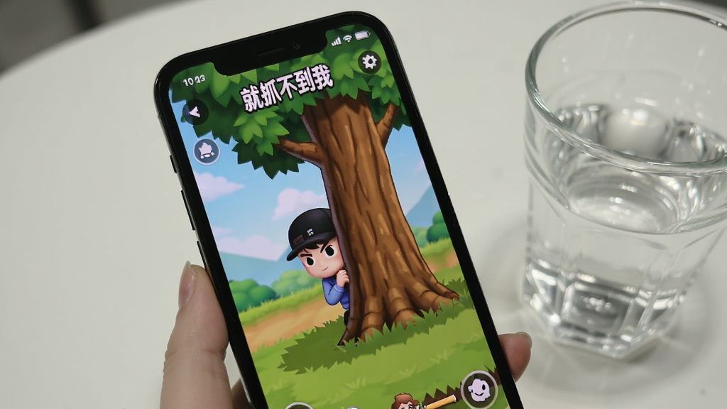 一张手机屏幕特写,屏幕上正在运行“就抓不到我”游戏,画面显示角色躲藏在树后,游戏界面清晰,手机旁边放着一杯水,背景简洁,突出使用场景,画质干净无多余元素。