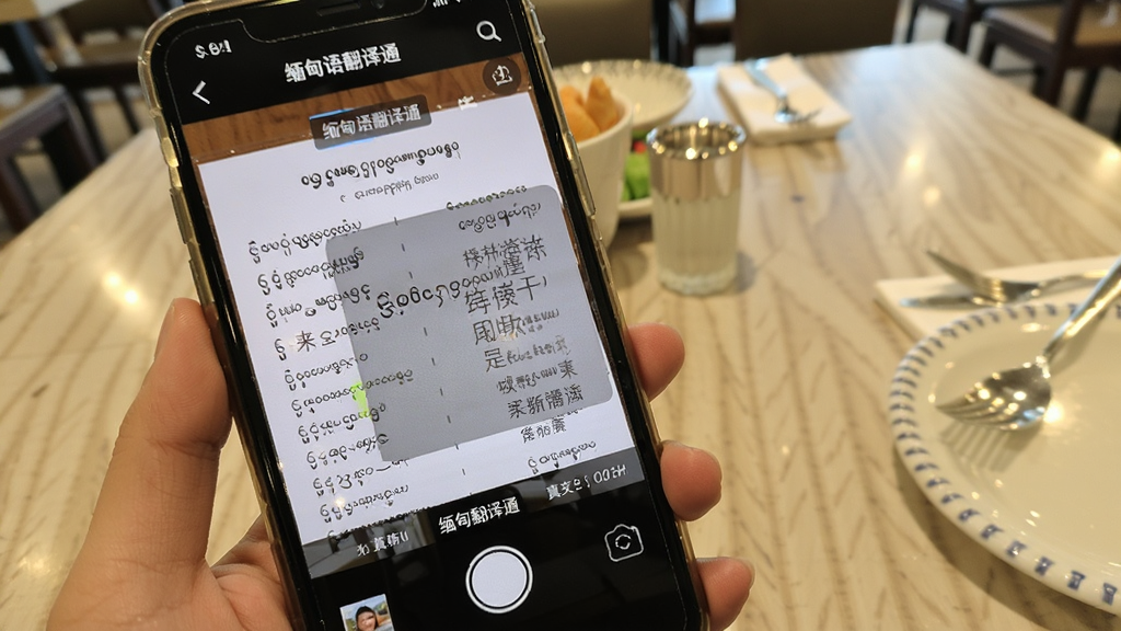 一部手机屏幕中显示“缅甸语翻译通”的拍照翻译界面,摄像头对准一份缅甸语菜单,屏幕上叠加了中文译文浮层,画面明亮清晰,背景是餐厅桌面,风格写实,画质干净,突出翻译工具的实用性
