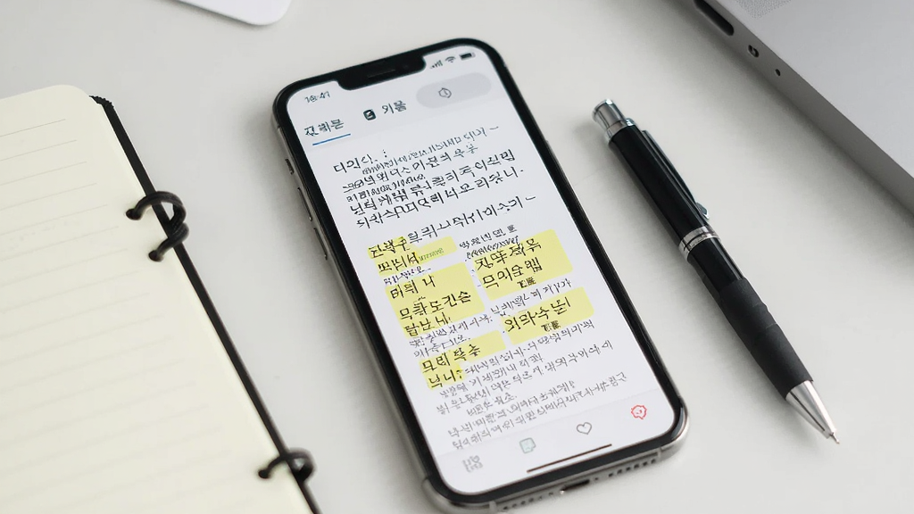 一张展示韩语翻译通术语翻译功能的界面图,画面中手机屏幕显示一份韩语技术文档的翻译结果,界面中高亮标注了专业术语的中韩对照,手机旁边放着一支笔和一本笔记本,背景是整洁的办公桌面,风格商务简洁,画质清晰,干净无多余元素