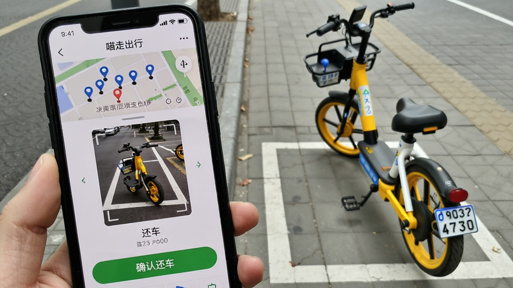 手机屏幕显示“喵走出行”App的还车界面,画面中有停车点地图、车辆停放拍照框和“确认还车”按钮,手机旁边有一辆黄色共享电单车停放在白色停车线内,背景为城市人行道,风格为写实教程截图风格,细节包括拍照提示文字、停车点标记、车辆车牌,画质清晰,干净无多余元素