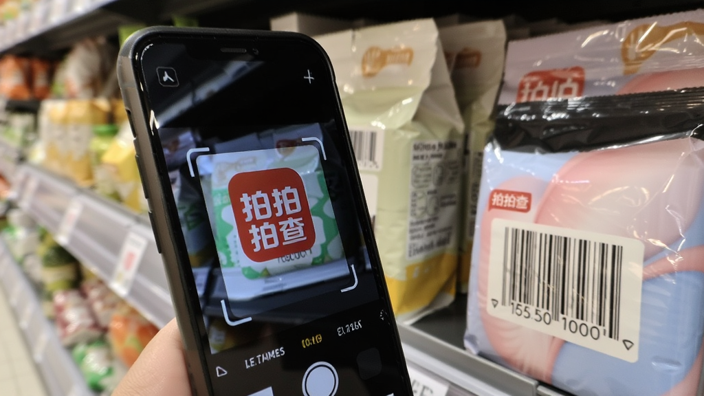主体是手机摄像头对准商品条形码进行扫描的场景,布局为手机在画面中央偏左、商品包装在右侧,排版为对角线构图,风格为写实生活风,细节包括手机屏幕显示扫描框和拍拍查软件logo、商品包装上的条码清晰可见、背景为超市货架虚化,画质清晰干净无多余元素