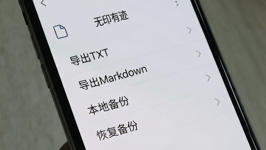 一张手机屏幕特写图,画面中显示“无印有迹”的导出菜单界面,菜单项包括“导出TXT”“导出Markdown”“本地备份”“恢复备份”,界面风格为浅灰色极简列表,文字清晰,布局整齐,画质干净,无多余装饰元素