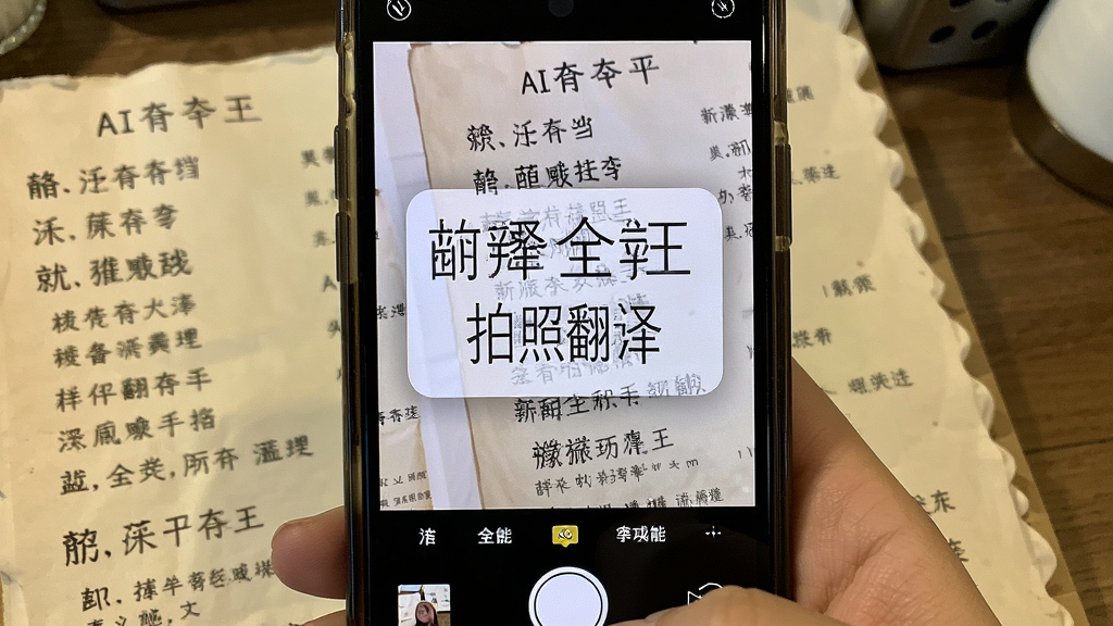 AI翻译全能王拍照翻译功能展示图,画面为手机摄像头对准一份外文菜单,屏幕上叠加显示翻译后的中文文字,布局为实景拍摄视角,风格真实生活化,细节包含菜单文字与翻译浮层,画质清晰干净无多余元素