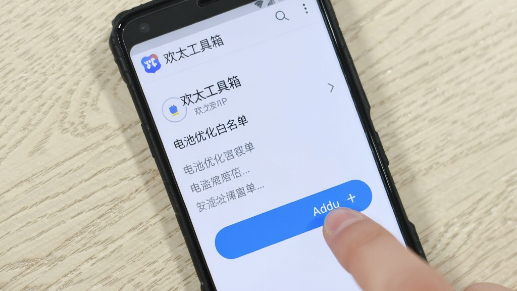 一部安卓手机屏幕显示“欢太工具箱”的电池优化白名单设置界面,手指正在点击添加按钮,背景为浅色木纹桌面,画面明亮清晰,风格生活化,画质1080P,干净无多余元素