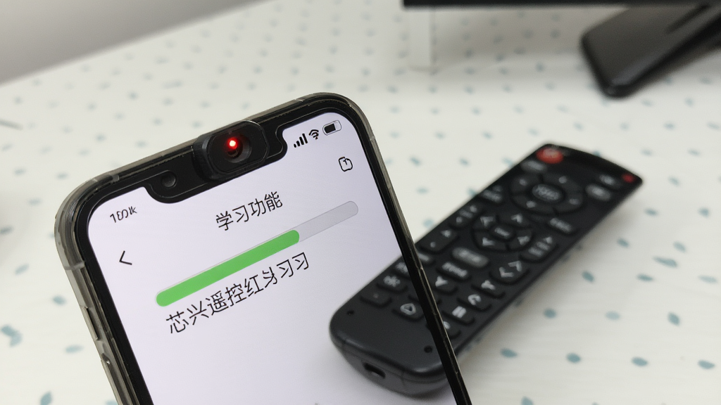 一张展示芯兴遥控红外学习功能的示意图,画面中一部手机顶部红外发射口对准一个传统遥控器,手机屏幕上显示学习进度条和提示文字,背景为简洁的桌面,风格写实,画质清晰,干净无多余元素
