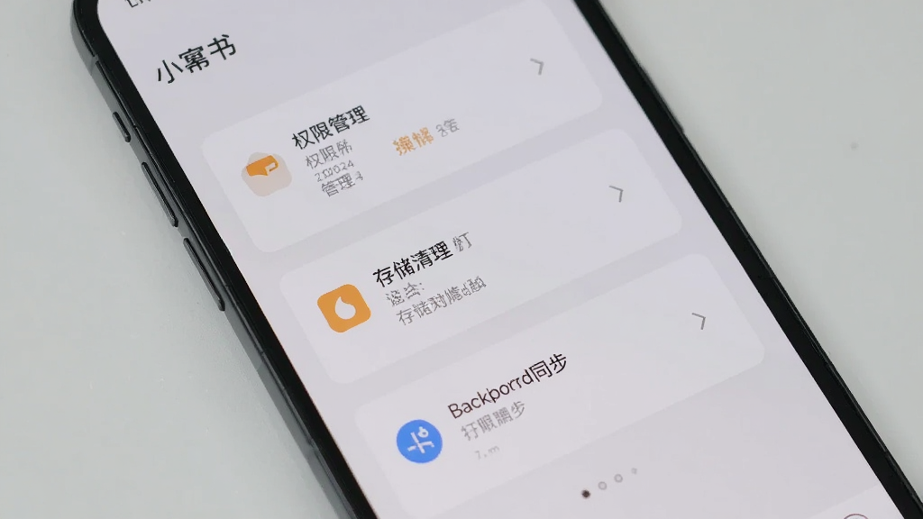 一部手机显示小幂书设置页面,包含权限管理、存储清理和后台同步选项,界面采用卡片式布局,配色柔和,背景为浅灰色,画面干净整洁,突出功能设置细节