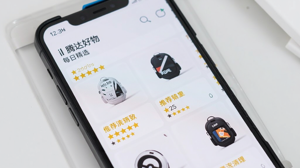 手机屏幕中展示“腾达好物”每日精选列表,商品图片排列整齐,旁边带有推荐标签与评分星标,背景为简洁的浅灰色桌面,风格清新明亮,画质清晰,无杂乱元素