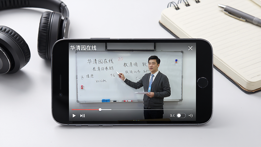 一张展示华清园在线视频播放界面的图片,画面中央是一部横屏放置的智能手机,手机屏幕上正在播放教学视频,视频画面中有一位老师在白板前讲课,屏幕下方有播放进度条和清晰度切换按钮,屏幕上方显示