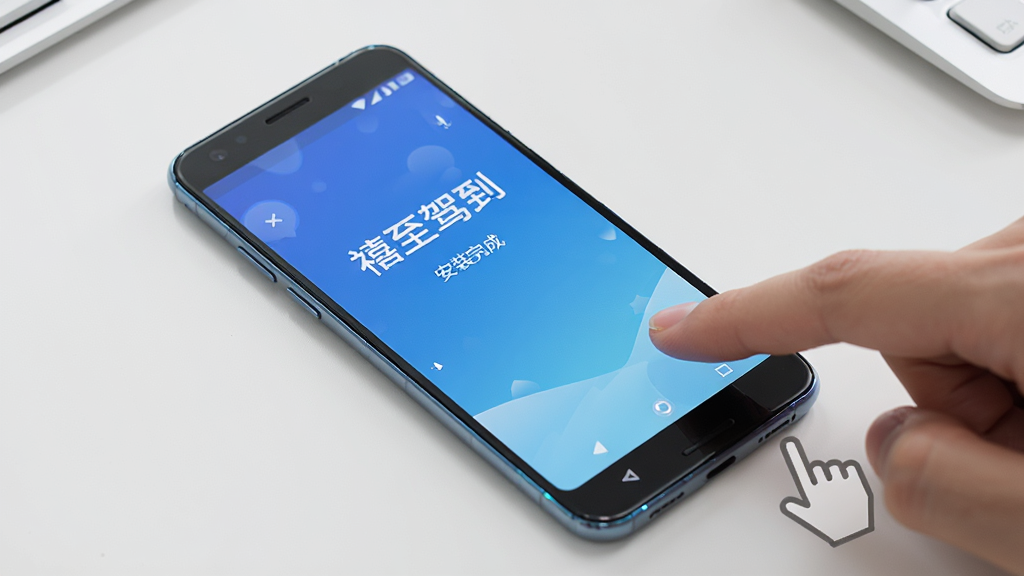 主体为一部安卓手机放置在桌面上,屏幕显示“禧至驾到”安装完成后的欢迎页面,旁边有手指点击屏幕的动作示意。布局为斜角俯视构图,风格为写实生活风,细节上手机边框有轻微反光、桌面干净整洁,画质高清干净,无多余元素。